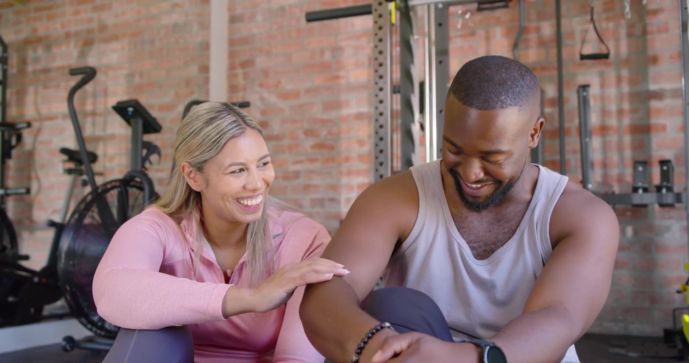 African American couple sitting in gym, woman in pink top reaching, touching his arm reassuringly