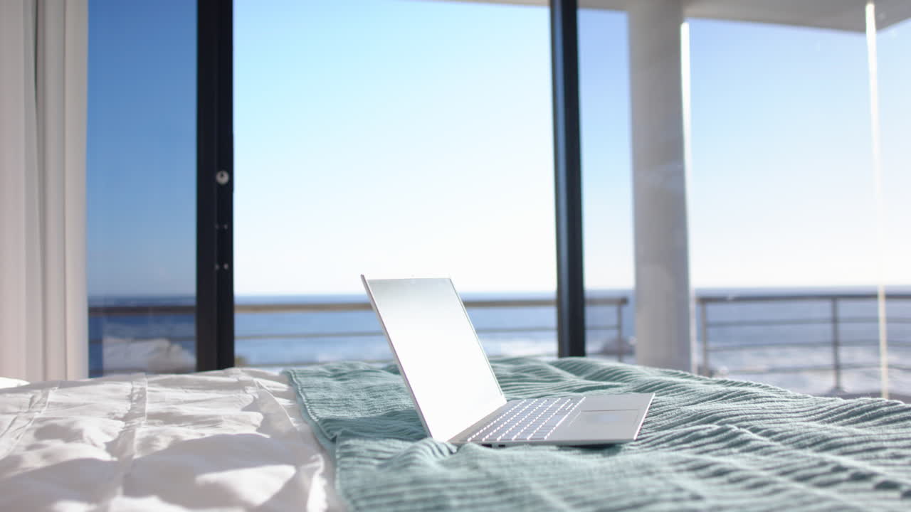 Laptop on bed with ocean view through large windows, creating serene workspace, copy space
