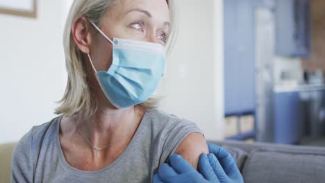 happy senior diverse woman and doctor wearing face masks in living room sitting on sofa, vaccinating