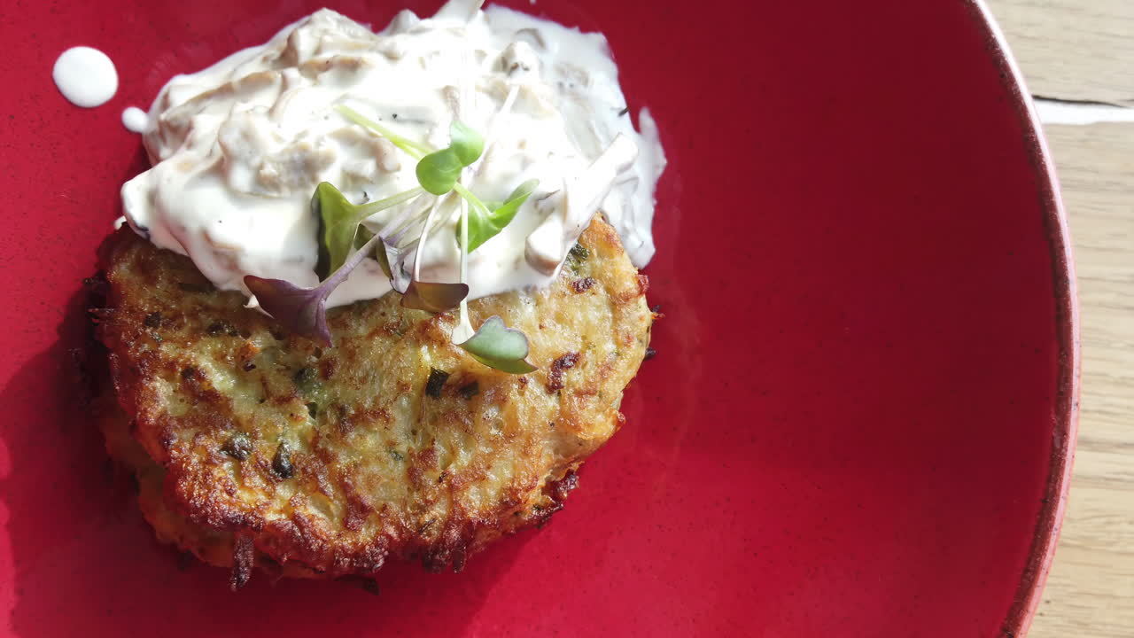 A golden-brown vegetable pancake sits on a red plate, topped with a smooth creamy sauce and fresh microgreens. Enjoy the delicious flavors and textures in this welcoming cafe setting