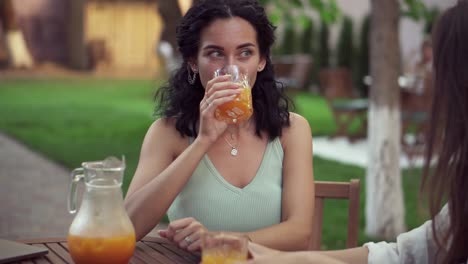 young women drinking orange juice and talking at outdoor cafe, smiling - portrait of brunette woman drinking juice