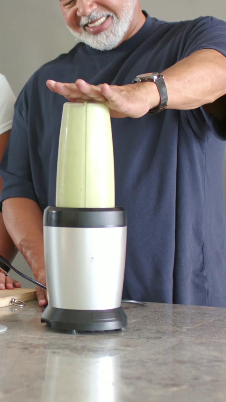 Video vertical: Hombre maduro con camiseta azul marino presionando hacia abajo, estabilizando una taza en la cocina, revisando la mezcla.
