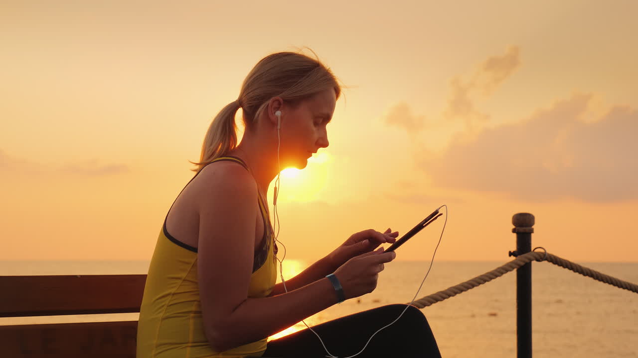 mujer fitness disfruta de una tableta sentada en un banco en un muelle contra el telón de fondo del sol naciente 4k vi