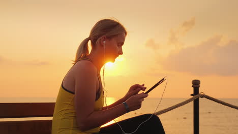 fitness woman enjoys a tablet sits on a bench on a pier against the backdrop of the rising sun 4k vi