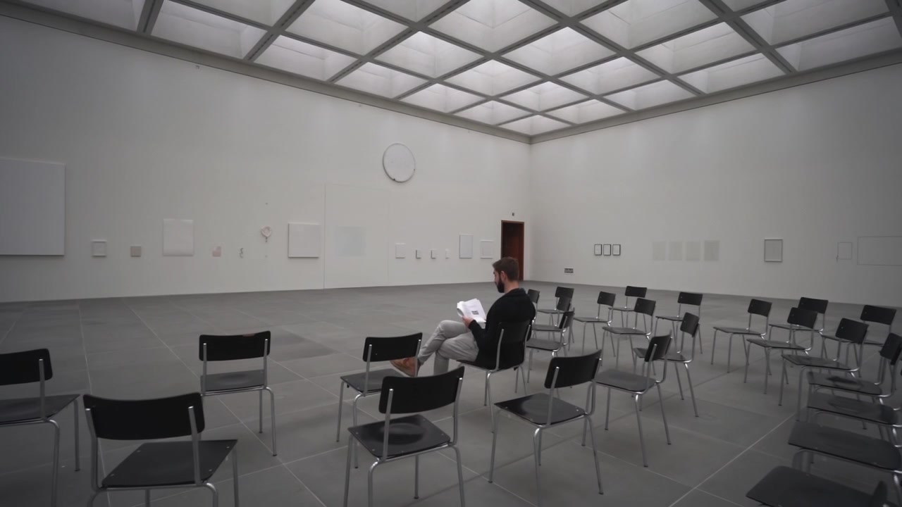 Giovane uomo solitario siede su una delle tante sedie in una spoglia sala bianca di museo e attende l'inizio di una presentazione