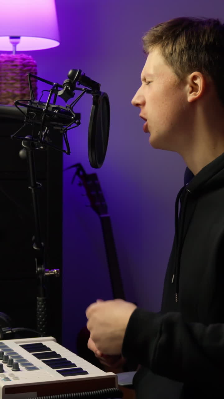 Mand set fra siden med hovedtelefoner på, der optræder foran en studiomikrofon med popfilter og gestikulerer, mens han optager i et lilla-belyst hjemmestudie med MIDI-keyboard og guitar i baggrunden