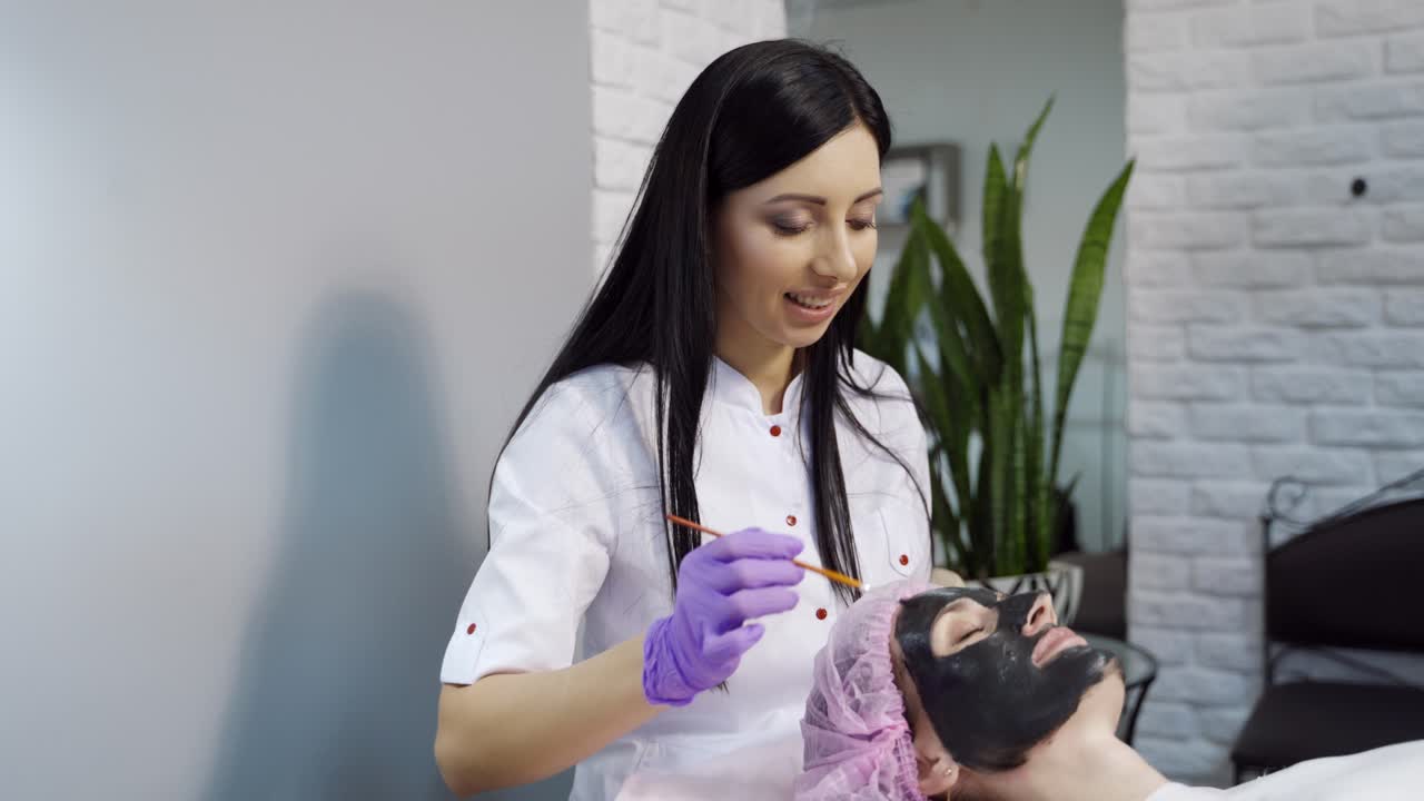 A beautiful cosmetologist applies a mask to clean the pores on the face of the client and communicates with her during the procedure at the beauty center.