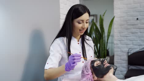 a beautiful cosmetologist applies a mask to clean the pores on the face of the client and communicates with her during the procedure at the beauty center.