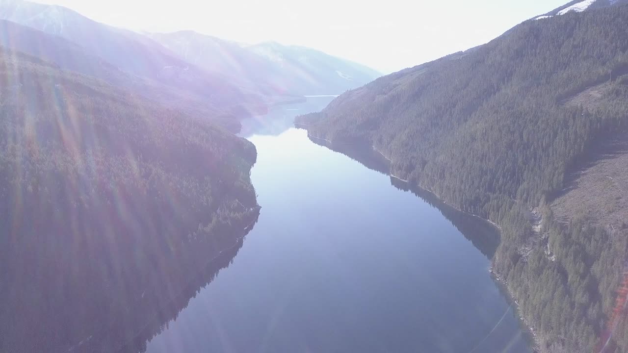 High aerial pan of forested deep blue mountain lake with lens flare