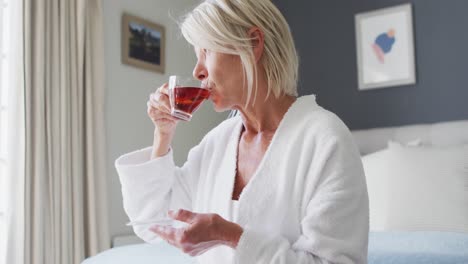 Happy-senior-caucasian-woman-sitting-on-bed-in-bedroom,-drinking-tea