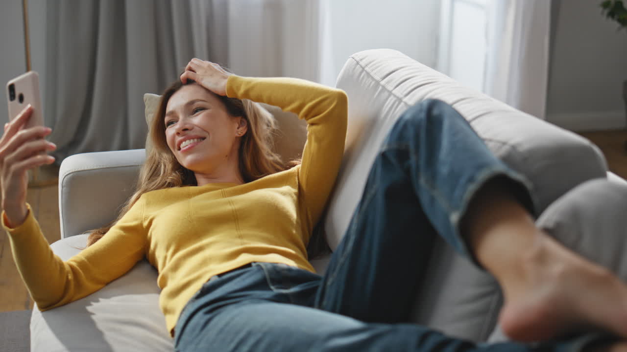 Friendly lady waving hand speaking smartphone at home. Relaxed woman laying sofa