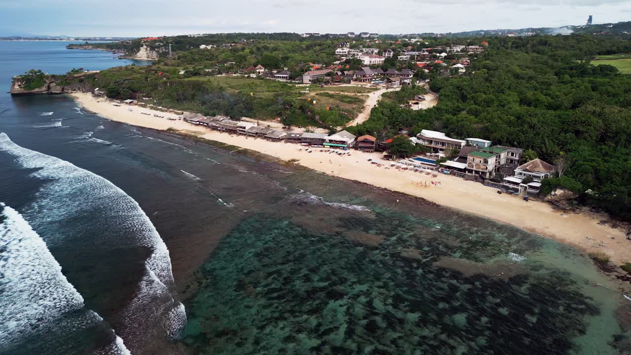 Drone footage capturing a broad sandy shoreline with shallow clear water, exposed coral reefs and a line of wooden huts along the coast forming a relaxed tropical setting under soft natural light