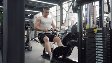 muscular man performs seated cable row on low row machine pulling heavy weight with full strength in busy modern gym environment while towel draped on bench and other gym goers move around