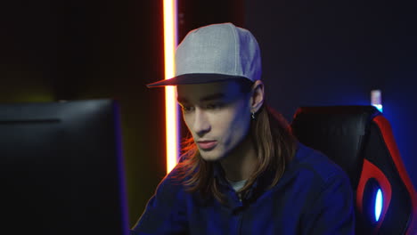 close up view of young serious man wearing cap and playing a game on the computer in a room with colorful neon lamps on the wall 2