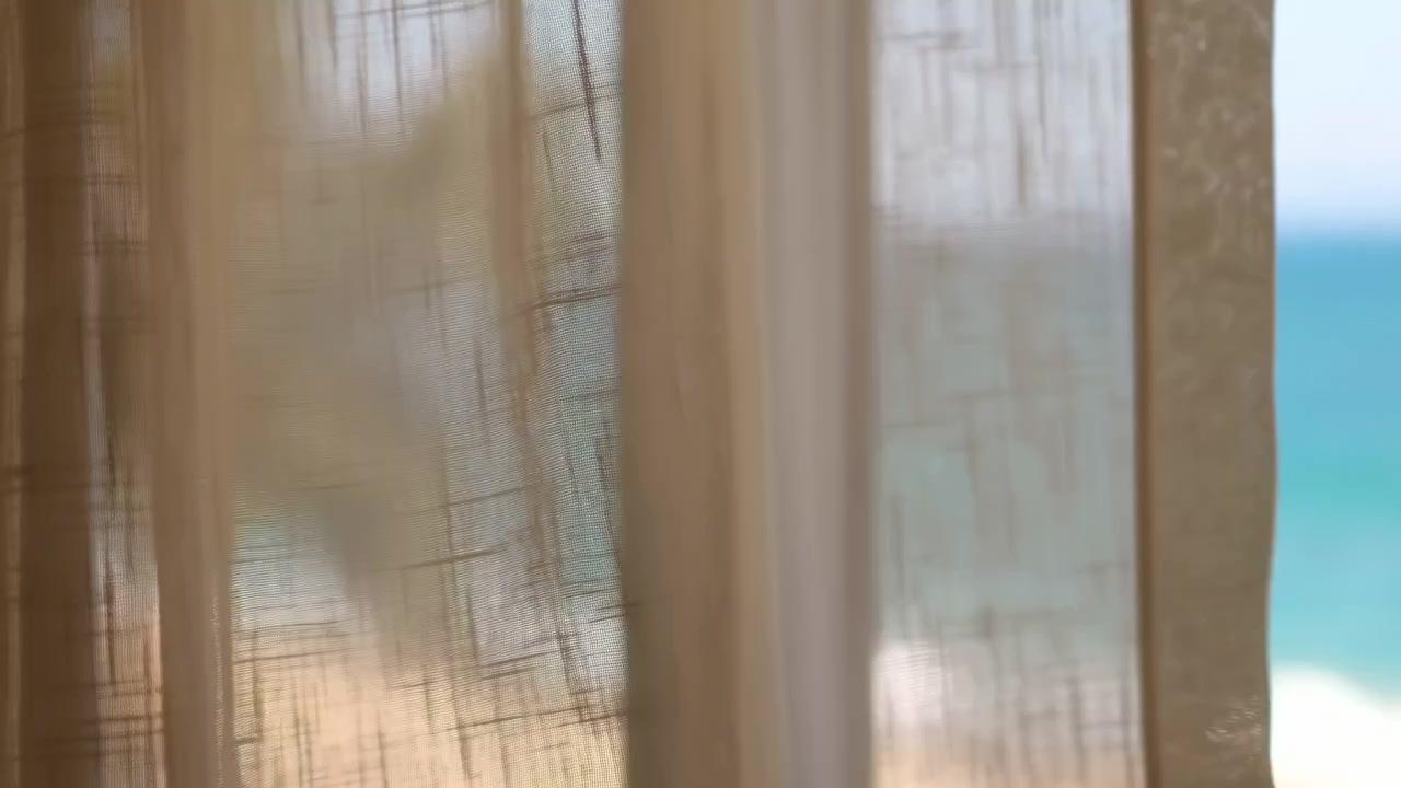 Translucent beige curtains hang near a sunlit window, with a softly blurred blue ocean visible beyond the glass. Gentle daylight filters through the fabric