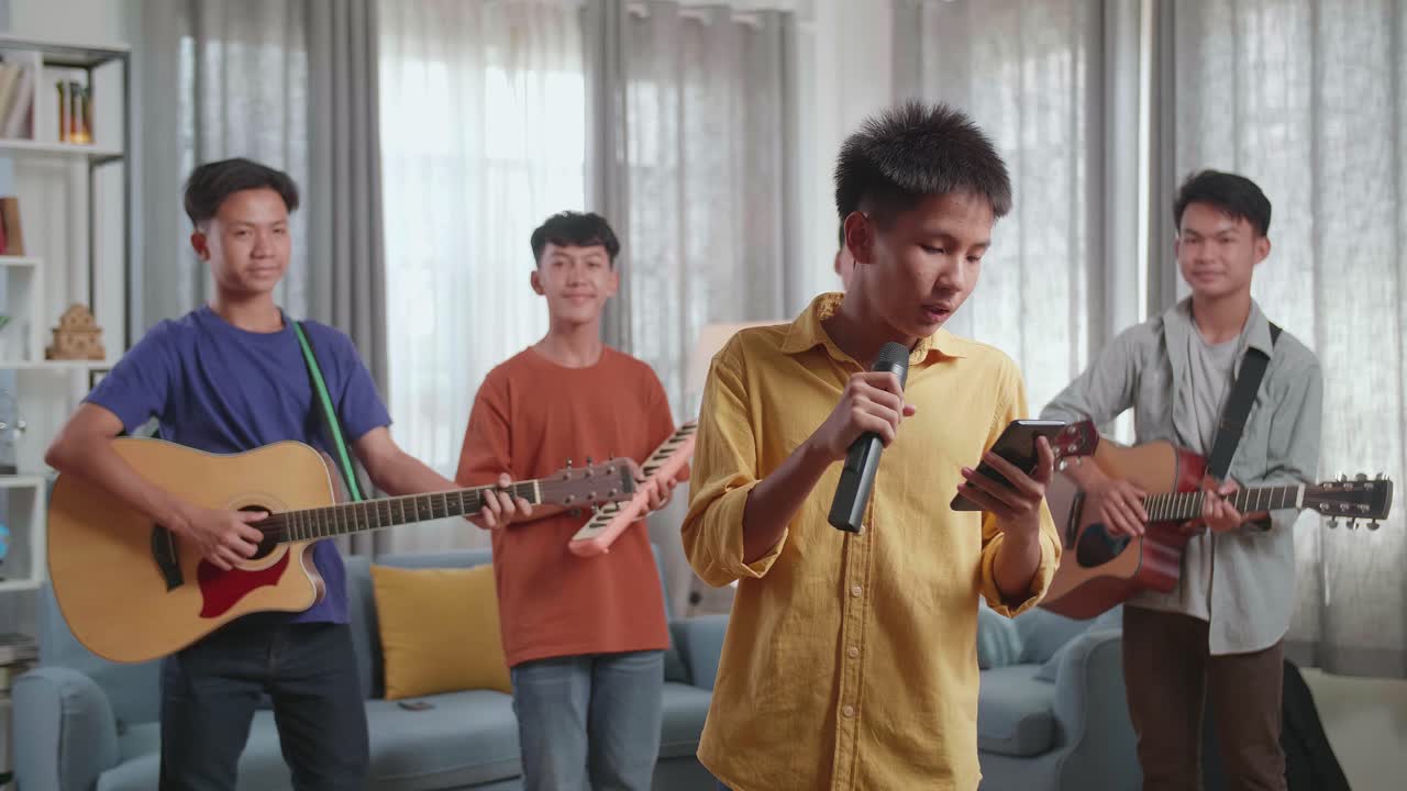 Young Asian Boy In Front Of His Friends With Musical Instruments Holding A Microphone And Looking At Script On Smartphone Before Speaking At Home