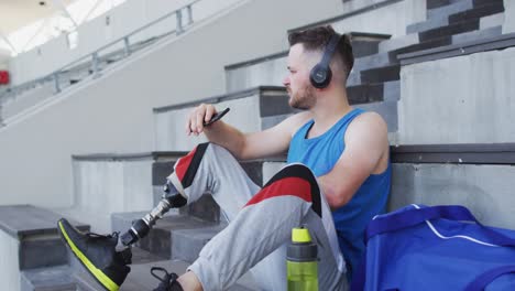 Caucasian-disabled-male-athlete-with-prosthetic-leg-sitting,-wearing-headphones,-using-smartphone