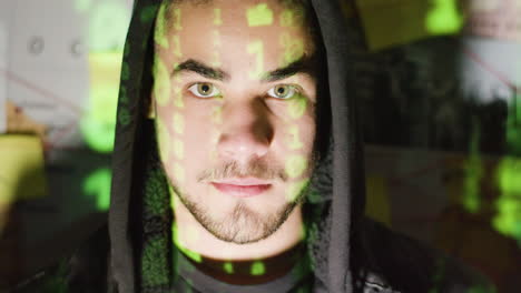 young male hacker in hood raising his head and looking at camera with binary code reflection on his face