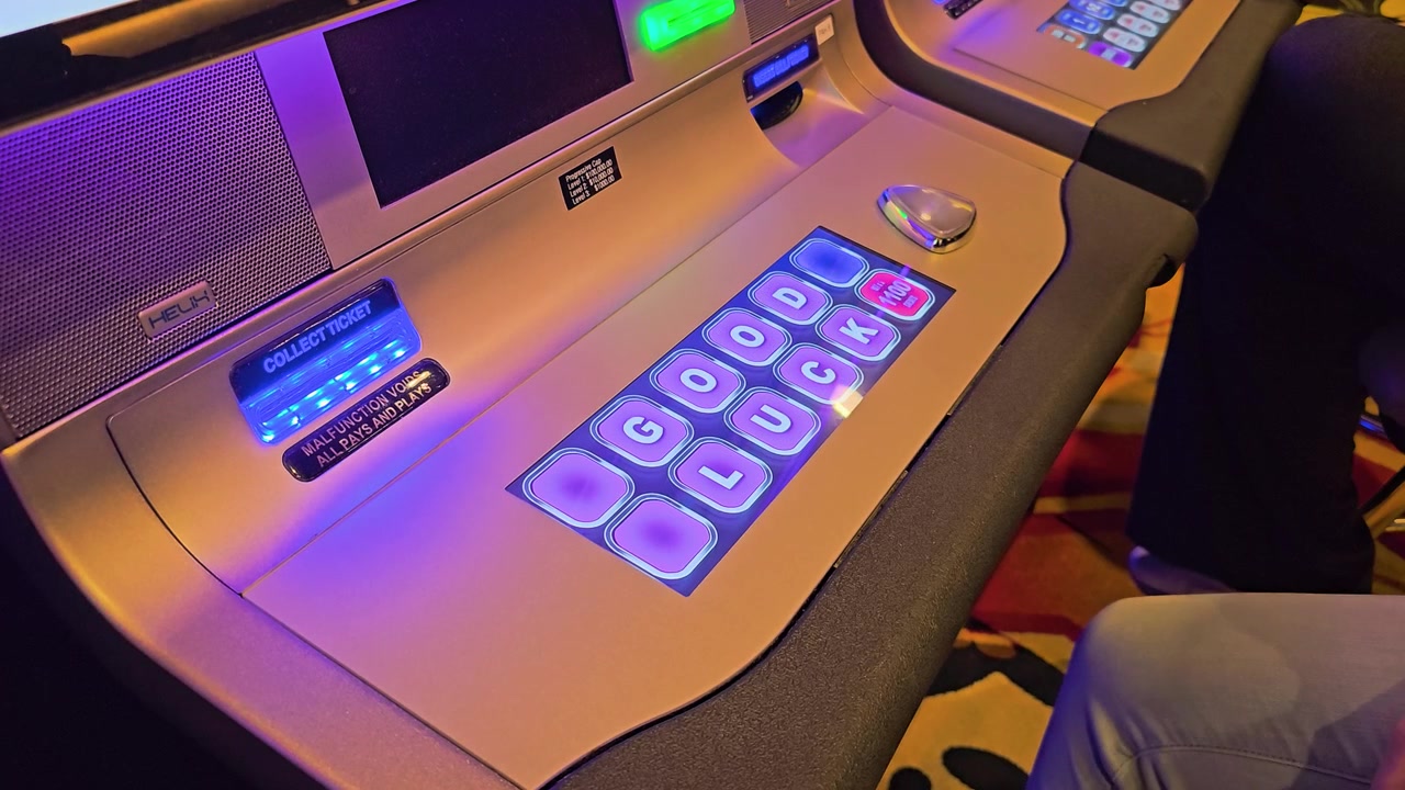 Casino player reaches for the metallic play button on a modern slot machine console, surrounded by glowing wager controls, ticket slot, colorful neon reflections, and seated gamblers nearby.