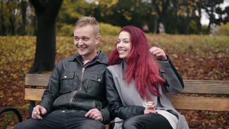 Young-attractive-couple-sitting-on-a-bench-in-autumn-park-and-talking.-Attractive-woman-with-red-hair-is-smoking