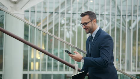 optimistic businessman walking upstairs, smiling while looking on smartphone