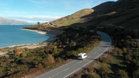 Drone-Aéreo-Siguiendo-Una-Autocaravana-Con-Vistas-Al-Hermoso-Lago-Azul-Hawea,-Campo-De-Ovejas-Y-Montañas-A-última-Hora-De-La-Tarde-En-Nueva-Zelanda