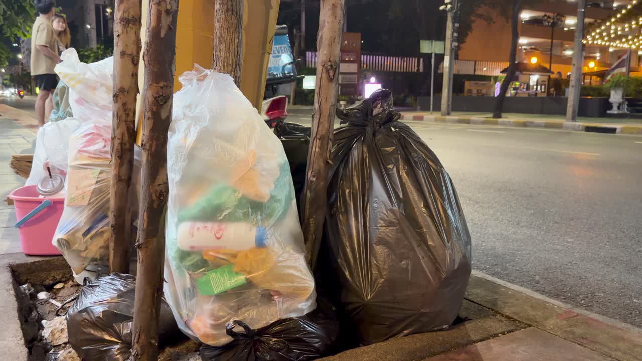 Garbage bags on sidewalk at night with vehicles and motorbike passing on busy Bangkok street