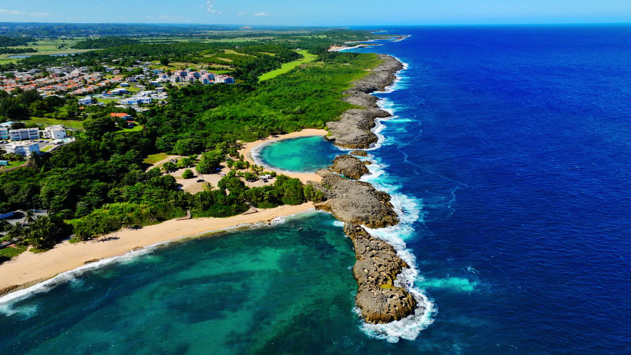 Vista dall'alto di una costa rocciosa con piscine naturali marine e una città costiera con edifici bianchi e campi verdi. Litorale e zona residenziale a Manati.