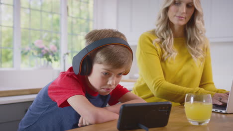 boy in kitchen watching movie on mobile phone wearing wireless headphones as mother works on laptop