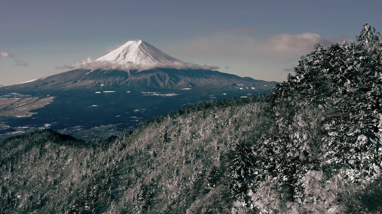 Majestic aerial footage of Japan’s iconic Mount Fuji, blanketed in pristine snow and partially veiled by drifting clouds. The foreground features a dense forest landscape