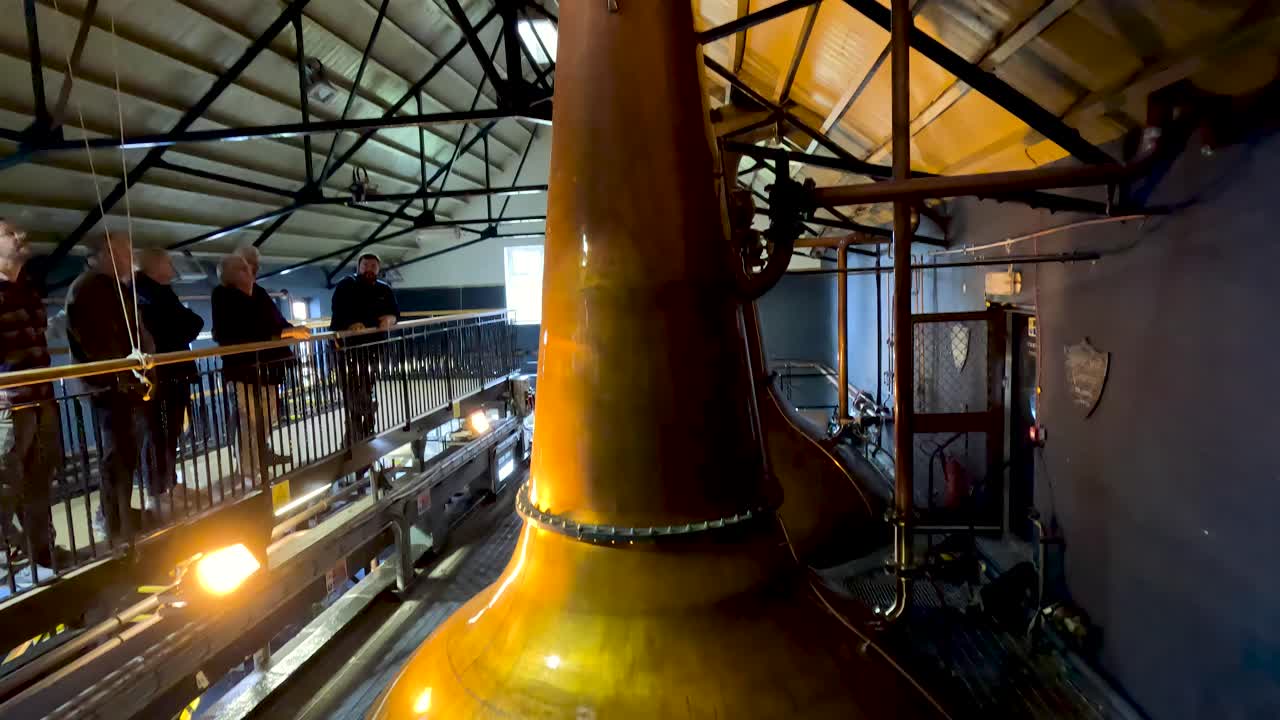 Wide-angle camera pans copper pot still, industrial distillery interior, warm lighting, visitors observing process