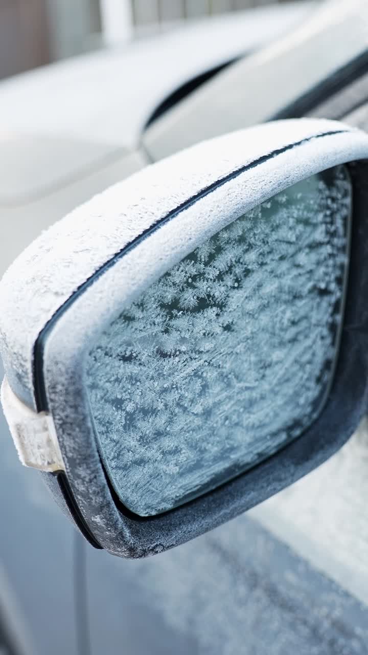 Primo piano dello specchietto laterale di un'auto ricoperto di brina e cristalli di ghiaccio in una fredda mattina invernale, che mostra la consistenza del vetro ghiacciato e le difficoltà di visibilità in condizioni climatiche di gelo