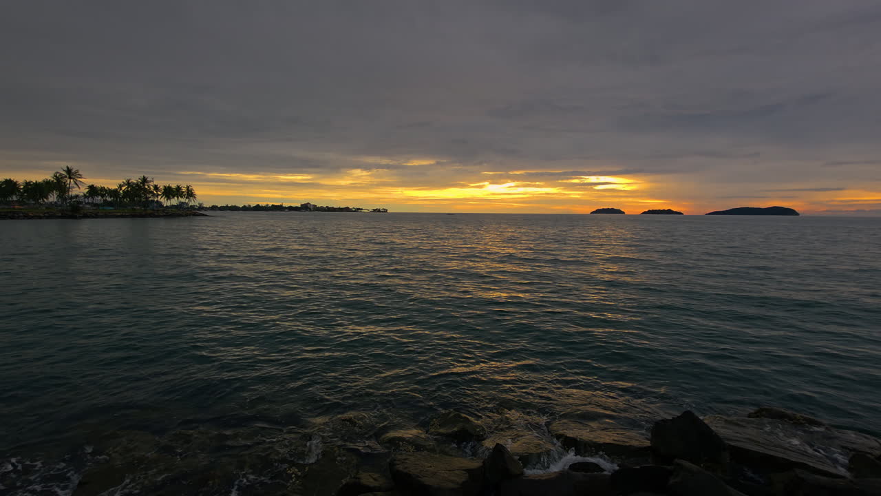 Kota Kinabalu, Sabah, Malaysia -
Golden Hues Across Cloudy Sunset Sky Over The Sea, Wave Crashing Against Rocky Beach
