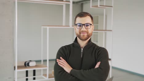 un hombre con un suéter y anteojos con barba está mirando a la cámara y sonriendo mientras cruza los brazos en una nueva oficina elegante con una apariencia caucásica