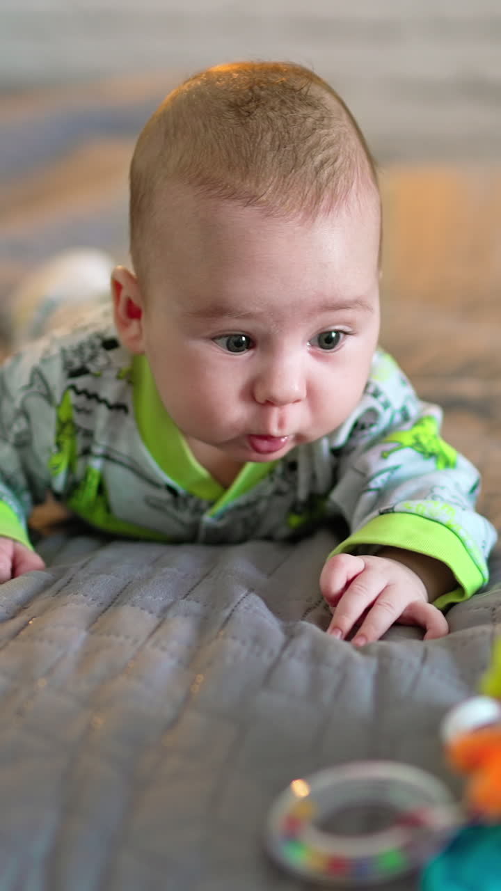 Tiny boy lies on the bed and toy is in front of him. Cute kid moving arms and legs actively, making funny faces. Grey backdrop in blur. Vertical video