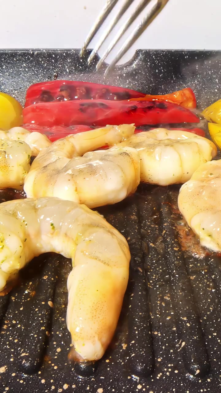 Inquadratura statica di una forchetta che gira peperoni rossi e gialli arrostiti su una padella grill antiaderente scanalata, accanto a gamberi pelati che sfrigolano su una superficie di granito scuro maculato