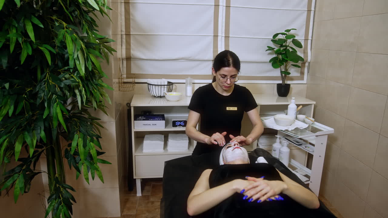 Female cosmetologist applies mask on the client's face. Beauty specialist carefully performs massage to a woman lying on the couch.