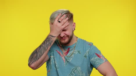 a young man with blonde hair and a beard wearing a floral shirt, a necklace, and tattoos. he is smiling and winking, looking at the camera. he is standing against a yellow background and gesturing with his hand.