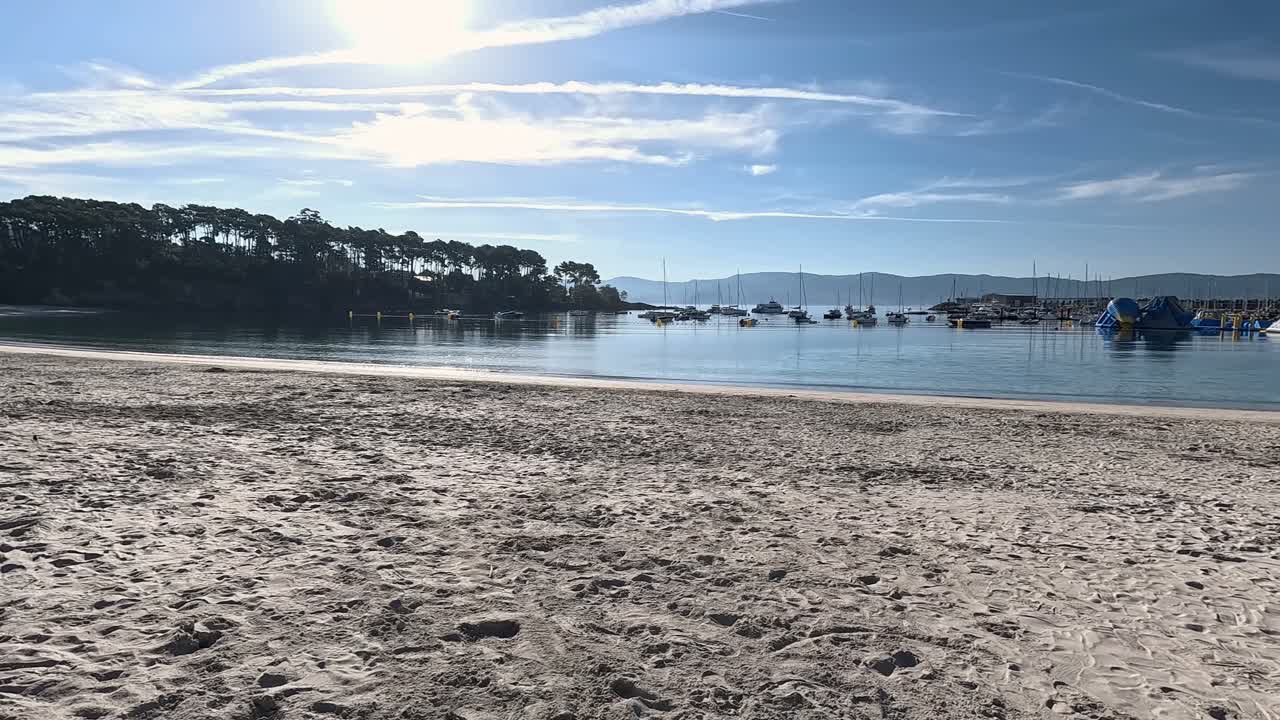 gente caminando a lo largo de la orilla de la playa con marea baja al amanecer, barcos amarrados en la marina cerca de los edificios de vivienda del pueblo, disparo descriptivo viajando hacia los lados