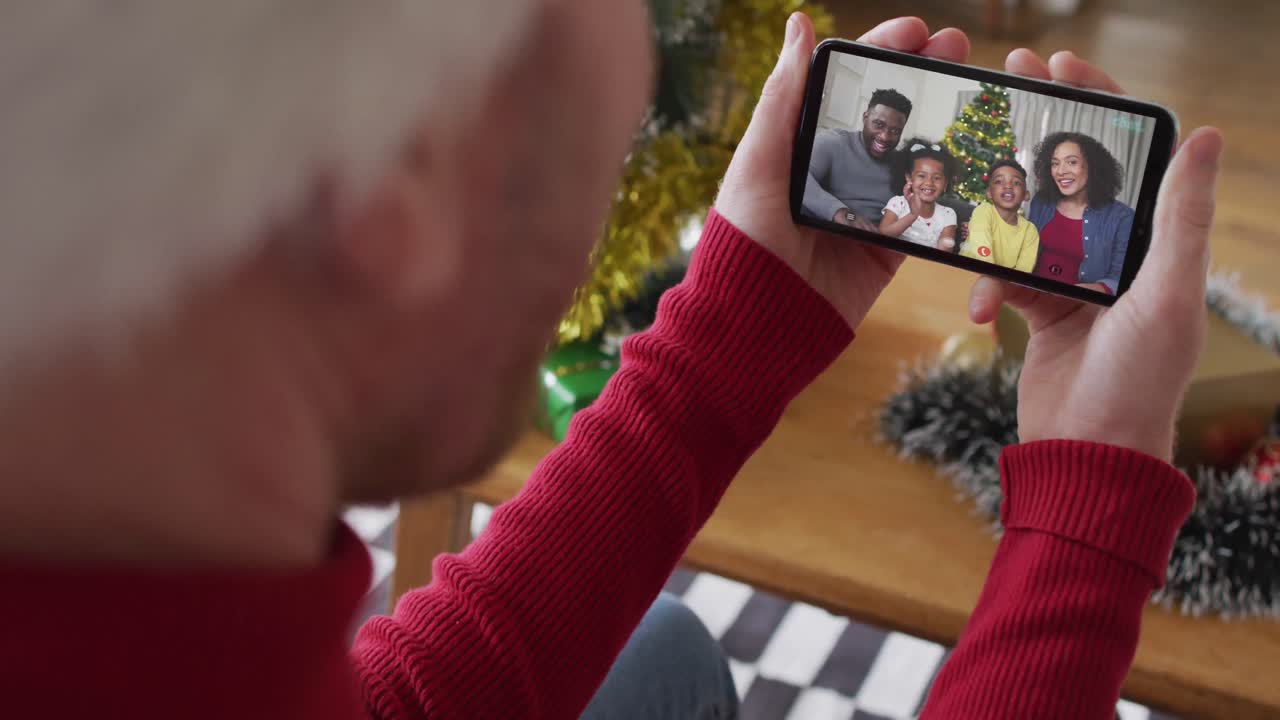 hombre caucásico con sombrero de santa usando un teléfono inteligente para una videollamada de navidad con una familia sonriente en la pantalla