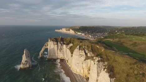 Volando-Sobre-El-Arco-Natural-De-Etretat-Con-La-Ciudad-Y-La-Bahía-Al-Fondo.