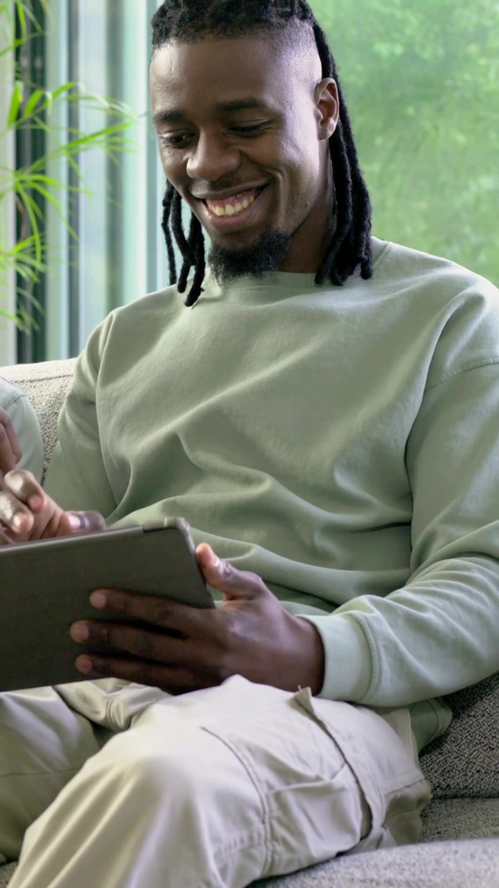 Vertical video: Tapping man in sweater using tablet by window woman in denim holding card showing