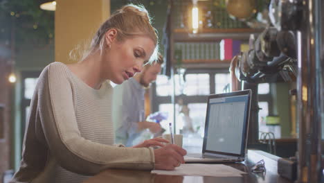 female manager of bar working at laptop on counter doing accounts with man cleaning in background