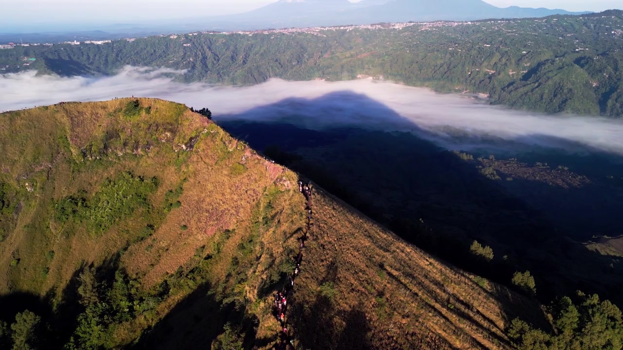 Escursionisti su cresta montuosa che camminano lungo un sentiero sopra un mare di nuvole in un paesaggio indonesiano ad alta quota con pendii ripidi e ampia vista sulle montagne nella soffusa luce mattutina, riprese aeree con drone