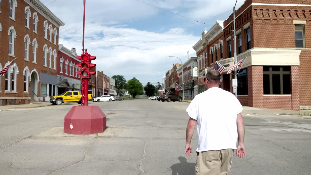 Man in white shirt and sunglasses walking towards an antique four way stop light in downtown Toledo, Iowa with gimbal video following from behind