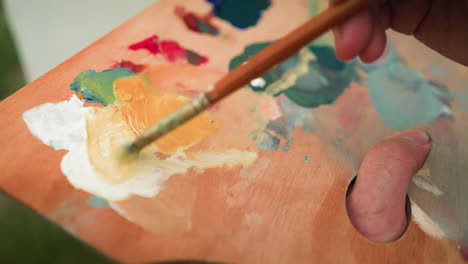 a close-up shot of an artist s hand holding a wooden palette while mixing various shades of paint with a small brush. the focus on the palette and brush