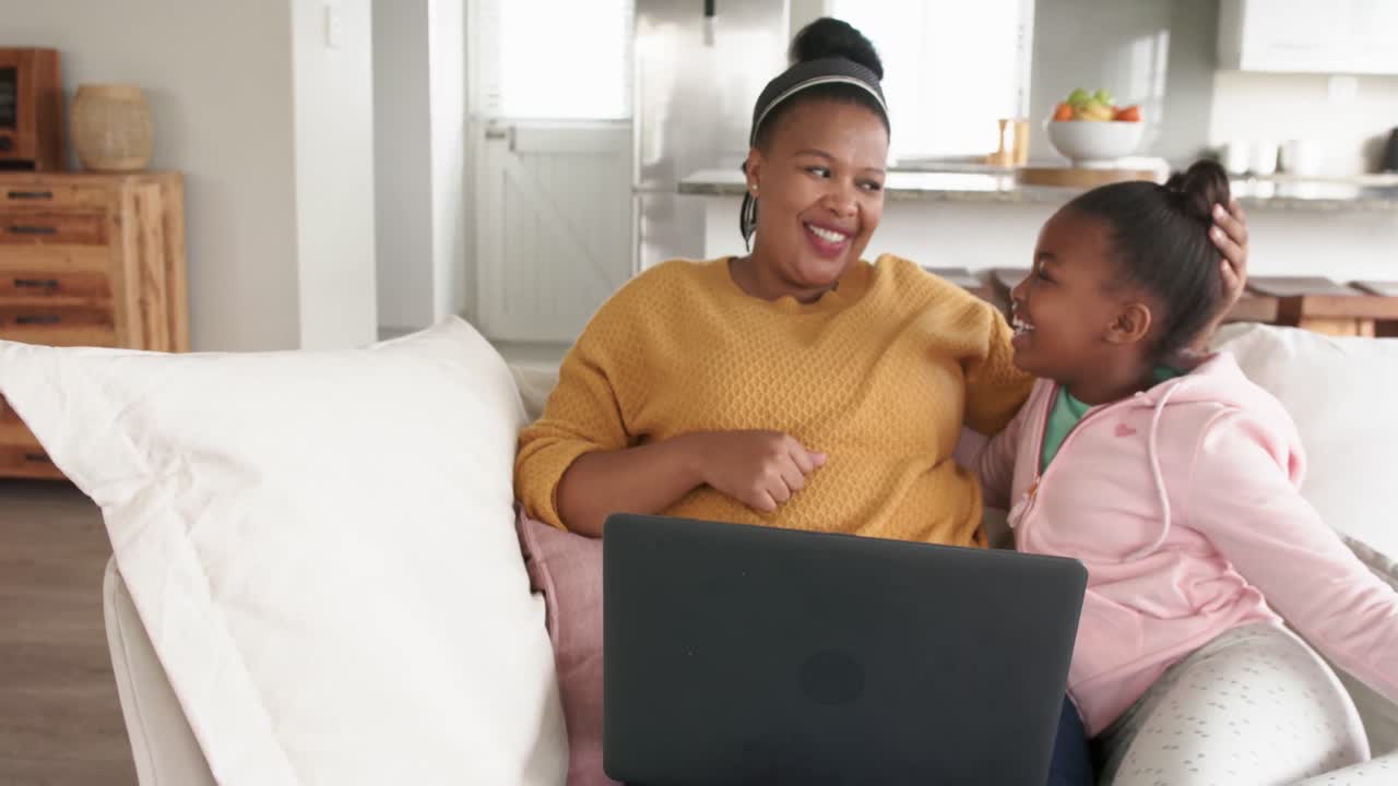 Happy african american mother and daughter relaxing on sofa using laptop, in slow motion