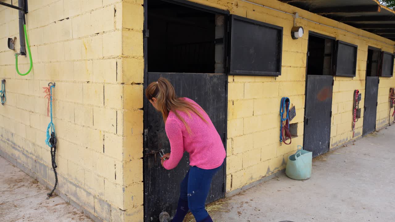 Young woman gives a bucket of food to her horse, then closes and latches the door, showing the daily feeding routine at a stable, equestrian center