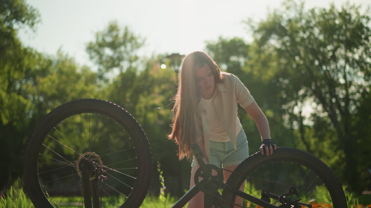 Young woman slightly bent adjusts front tire of her upside-down bike while rotating back tire, hair flowing down, sunlight reflecting off her, surrounded by lush greenery, background includes trees