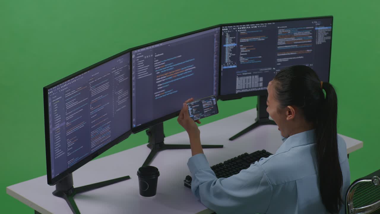 vista lateral de una desarrolladora asiática feliz mirando el código en un teléfono móvil mientras escribe código con múltiples pantallas de computadora en el estudio de pantalla verde